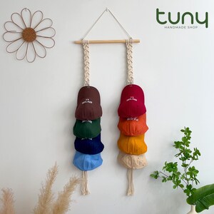 Peut inclure: Un organisateur de chapeaux en macramé avec une cheville en bois et deux colonnes suspendues. Chaque colonne contient une pile de casquettes de baseball colorées, dont marron, rouge, vert, bleu, orange, jaune et beige. Les casquettes portent les inscriptions "New York" et "Paris".