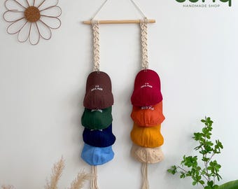 Könnte beinhalten: Ein Makramee-Hut-Organizer mit einem Holzdübel und zwei hängenden Säulen. Jede Säule hält einen Stapel bunter Baseballkappen, darunter braun, rot, grün, blau, orange, gelb und beige. Die Kappen tragen die Aufschriften "New York" und "Paris".