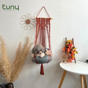 May include: A handmade macrame pet bed in rust-colored cord, suspended from a wooden dowel. A small, gray dog rests comfortably in the bed, which is a soft, round, gray cushion. The Etsy shop logo "tuny handmade shop" is in the upper left corner.