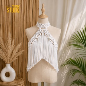 Op de afbeelding: Een wit macramé haltertop met een hoge halskraag, ingewikkelde geknoopte geometrische patronen op de borst en lange, vloeiende franjes die vanaf de zoom naar beneden hangen. Het kledingstuk wordt getoond op een beige paspop.
