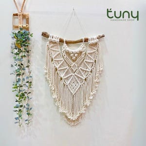 Peut inclure: Une tenture murale en macramé crème avec un motif géométrique, suspendue à une branche en bois naturel. La pièce faite à la main présente des motifs de nœuds complexes et un bas à franges. La décoration murale est exposée sur un mur blanc.
