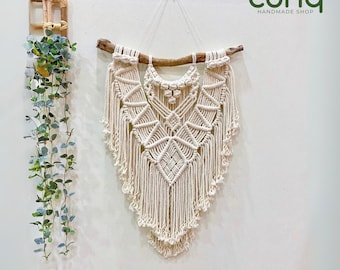 May include: A cream-colored macrame wall hanging with a geometric design, suspended from a natural wooden branch. The handmade piece features intricate knotting patterns and a fringed bottom. The wall decor is displayed against a white wall.