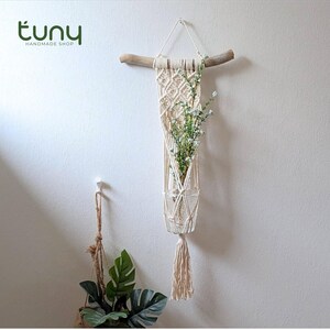 May include: A cream-coloured macrame plant holder with a glass vase, suspended from a natural wood branch. The woven design holds a clear glass vase filled with greenery and small white flowers. The bottom of the holder has a tassel. The shop name "tuny HANDMADE SHOP" is in the top left.