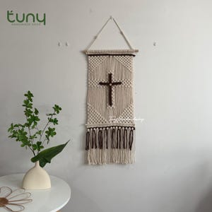May include: A rectangular macrame wall hanging featuring a central dark brown cross motif against a cream-colored cord background. The piece is suspended from a wooden dowel with a triangular cord hanger and finished with a fringe of alternating cream and dark brown tassels at the bottom edge.