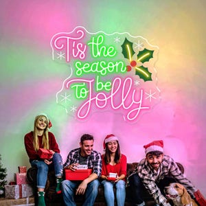 May include: A neon sign with the text "Tis the season to be Jolly" in pink and green, with holly and snowflake accents. Four people are sitting on a couch, holding gifts, and wearing Santa hats. A dog is also present.