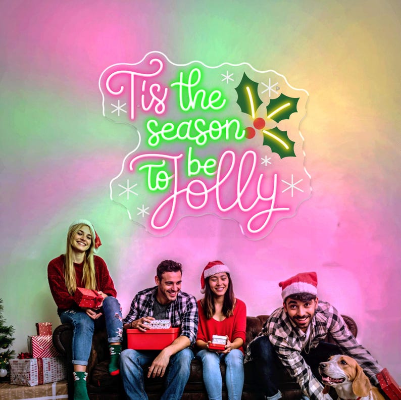 May include: A neon sign with the text "Tis the season to be Jolly" in pink and green, with holly and snowflake accents. Four people are sitting on a couch, holding gifts, and wearing Santa hats. A dog is also present.