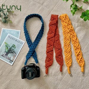 May include: Macrame camera straps in teal, rust, and yellow. A black Canon camera is attached to the teal strap. The straps are displayed on a textured, neutral-coloured surface, with a magazine and greenery in the background.