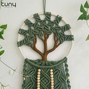May include: A handmade macrame wall hanging featuring a tree design. The tree has a brown trunk and olive green leaves, set within a cream-colored circular frame. The bottom section includes olive green macrame with wooden beads.