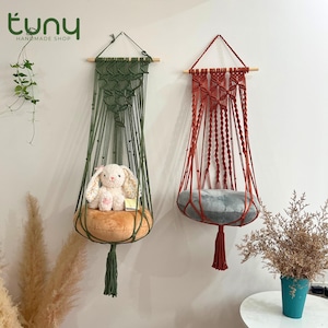 May include: Two macrame hanging planters, one green and one red, suspended from wooden dowels. The green planter holds a plush bunny on a tan cushion, while the red one has a gray cushion. Both have a tassel at the bottom. The shop name "tuny handmade shop" is in the top left.