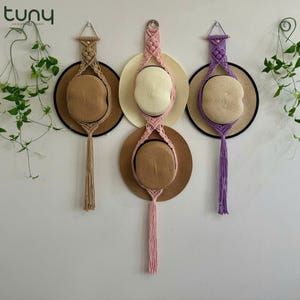 Puede incluir: Tres colgadores de sombreros de macramé en beige, rosa y morado, cada uno con un sombrero de paja. Los sombreros tienen bordes negros. Los colgadores de macramé están suspendidos de la pared, con una larga borla.