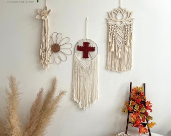May include: A collection of handmade macrame wall hangings in cream and natural tones. The designs include a flower, a daisy, a cross, and a lotus flower. The arrangement is displayed on a white wall, with a small wooden ladder and decorative leaves.