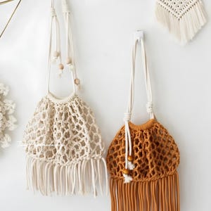 May include: Two macrame drawstring bags with knotted net patterns and long fringe at the base. One bag is off-white with light wooden beads on the cord, and the other is mustard yellow with matching wooden beads. Both feature long shoulder straps.