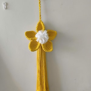 May include: A yellow macrame wall hanging featuring a five-petaled flower design with a white pom-pom center. The flower is attached to a braided cord that hangs from a light-colored wooden ring, with a long yellow fringe extending downward.