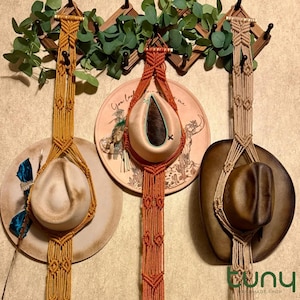Puede incluir: Tres colgadores de sombreros de macramé en tonos dorados, naranjas y beige, cada uno sosteniendo un sombrero de vaquero. Los colgadores están sujetos a un estante de madera con vegetación. Los sombreros se exhiben contra platos decorativos.
