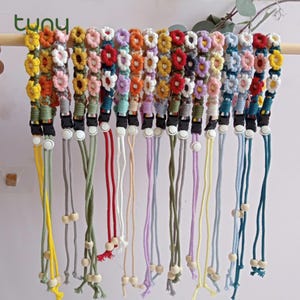 May include: A collection of colorful, daisy-shaped mask lanyards. Each lanyard features a different color daisy chain, with matching cords and wooden beads. The lanyards are hanging on a wooden dowel against a white background.