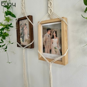 May include: Two wooden picture frames with white rope hangers, each displaying a photo of a couple. The frames are hanging on a white wall with green plants in the background.