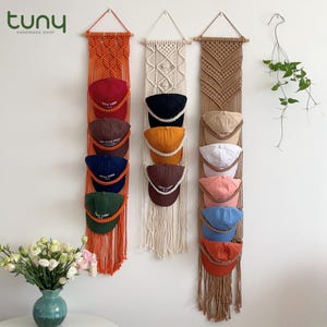 Pode incluir: Organizadores de chapéus em macramé laranja, creme e castanho, cada um com vários bonés de beisebol em várias cores. Os bonés apresentam o texto "NEW YORK". O logotipo "Tuny Handmade Shop" está no canto superior esquerdo.