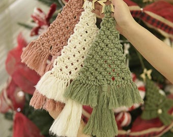 Könnte beinhalten: Drei handgefertigte Makramee-Weihnachtsbaumschmuck in Rosa, Creme und Grün, jeweils mit einem Holzstern versehen. Die Ornamente haben einen Fransenboden und werden vor einem geschmückten Weihnachtsbaum gehalten.