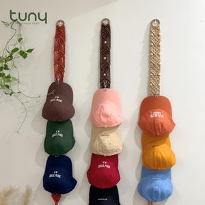 May include: Three macrame hat holders displaying baseball caps in diverse colours. The caps include brown, green, and navy blue with "N.Y." text, pink, beige, and red with "NEW YORK" text, and orange, yellow, and blue caps with "PARIS" and "CRUISE" text.