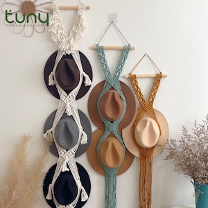 May include: Macrame hat holders in cream, teal, and tan, each with a wooden dowel and hanging rope. The holders display various fedora hats in brown, gray, and black. The wall decor adds a bohemian touch to any space.
