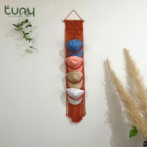 Peut inclure: Un organisateur de chapeaux en macramé orange brûlé avec cinq casquettes de baseball bleues, roses, beiges et blanches. Suspendu à une cheville en bois, il présente une base à franges. Le logo de la boutique Etsy est visible en haut à gauche.