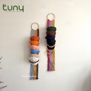 May include: Two macrame wall hangings featuring circular wooden rings at the top, designed to hold multiple baseball caps. The left organizer displays caps in shades of orange, pink, blue, and white, while the right holds dark-colored caps.