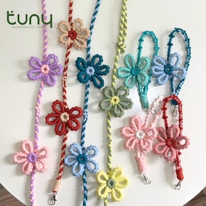 May include: A collection of colorful macrame flower lanyards. Each lanyard features a different colored flower, including pink, purple, blue, green, and yellow. The lanyards are handmade and have a small metal clasp.