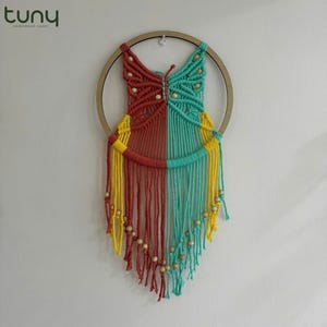 May include: A macrame wall hanging with a butterfly design in red, yellow, and turquoise. The butterfly is made of knotted string and has wooden beads around the edges. The hanging is attached to a wooden hoop.