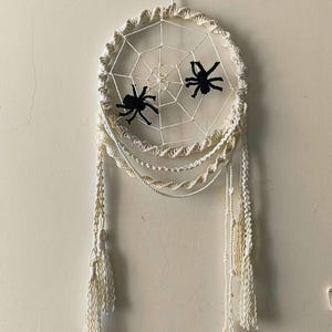 May include: A cream-colored macrame wall hanging featuring a circular frame with a woven spider web pattern. Two black spiders are attached to the web. The piece includes hanging cords with twisted details and long, fringed tassels at the bottom.