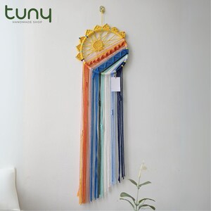 May include: A handmade dreamcatcher with a yellow sun-like design and colorful yarn strands. The dreamcatcher features orange, blue, white, and green yarn, with a small white tag hanging from the bottom. The shop name "tuny handmade shop" is in the top left corner.