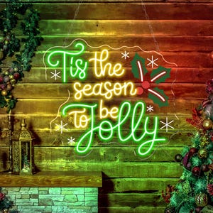 May include: A neon sign with the phrase "Tis the season to be Jolly" in green and yellow script. The sign includes holly and snowflake accents, hanging against a wooden background. A Christmas tree and decorative lantern are visible.