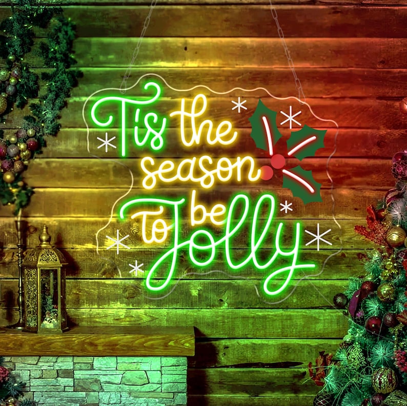 May include: A neon sign with the phrase "Tis the season to be Jolly" in green and yellow script. The sign includes holly and snowflake accents, hanging against a wooden background. A Christmas tree and decorative lantern are visible.