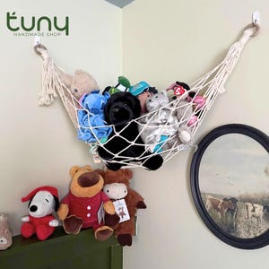 May include: A macrame hammock filled with plush toys, suspended in a corner. The hammock is cream-colored and holds a variety of stuffed animals, including a black dog, a blue character, and several others. Below, several more plush toys sit on a green shelf.
