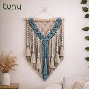 May include: A macrame wall hanging with a wooden dowel, featuring a geometric design in blue, beige, and gray. The piece includes tassels and wooden beads, creating a bohemian aesthetic. The text "tuny handmade shop" is in the upper left corner.