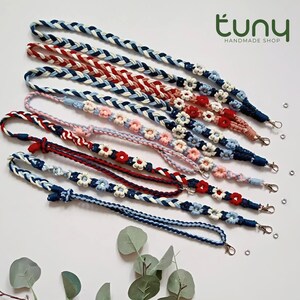 May include: A collection of handmade lanyards in various colors, including blue, red, and pink. Some lanyards feature a braided design, while others are adorned with small flower accents. Each lanyard has a metal clasp.