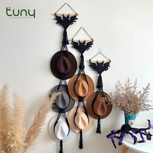 May include: A macrame hat rack with three tiers, each featuring a black bat design and wooden dowel. The rack holds various fedora hats in shades of brown, gray, and white. The Etsy shop logo "tuny handmade shop" is visible in the top left corner.