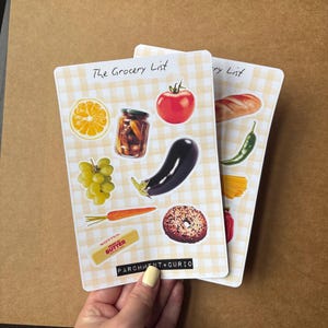 以下が含まれることがあります： 黄色のギンガムチェック柄の買い物リストのメモ帳が2冊。各メモ帳には、トマト、ナス、ベーグルなどの食品のイラストが描かれています。「The Grocery List」の文字がメモ帳の上部に印刷されています。「PARCHMENT+CURIO」のブランド名が下部に印刷されています。