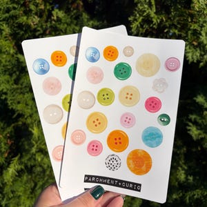 Pode incluir: Dois cartões brancos com ilustrações em aquarela de botões coloridos em vários tamanhos e cores, incluindo azul, laranja, verde, rosa e amarelo. O texto "PARCHMENT+CURIO" está impresso na parte inferior dos cartões.