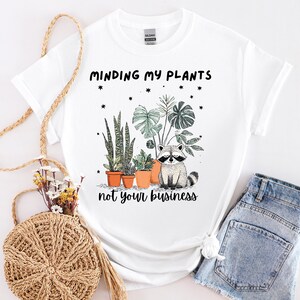 May include: White t-shirt featuring the text "Minding my plants not your business" and a graphic of a raccoon with potted plants. The plants include snake plants and monstera leaves. The shirt is styled with denim shorts.