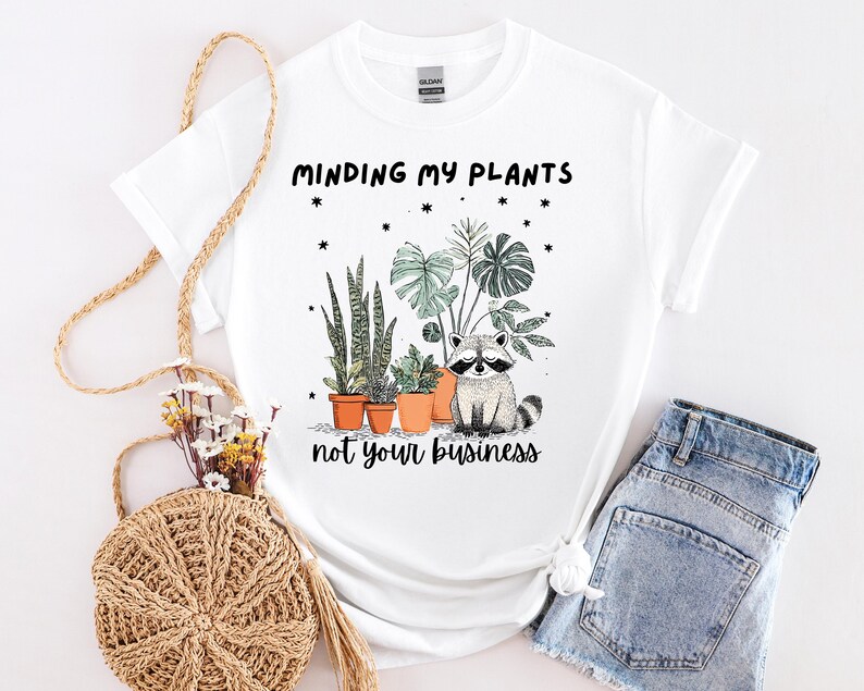 May include: White t-shirt featuring the text "Minding my plants not your business" and a graphic of a raccoon with potted plants. The plants include snake plants and monstera leaves. The shirt is styled with denim shorts.