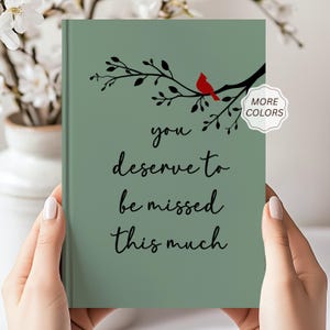 May include: A sage green hardback journal featuring a black silhouette of a tree branch with a small red cardinal bird perched on it. The text "you deserve to be missed this much" is printed in black cursive script in the centre of the cover.