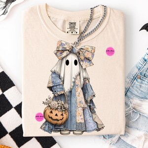 Op de afbeelding: Crèmekleurig T-shirt met een grillige spookillustratie in een patchwork denim en bloemenjurk, met een pompoenmand. De geest heeft een grote strik en twee zwarte ogen. Een zilveren kettingketting is over het shirt gedrapeerd. Het shirt heeft het Comfort Colors-merklabel.