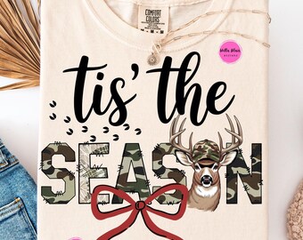 May include: Cream-colored t-shirt with the phrase "tis' the SEASON" in black script and camouflage print. A deer wearing a camouflage hat is featured, along with a red bow. A gold necklace and a Comfort Colors tag are visible.