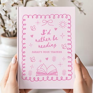 May include: A light pink book with a dark pink border and the text I'd rather be reading and SARAH'S BOOK TRACKER. The cover features line art illustrations of a bow, sun, hearts, flowers, butterflies, and an open book in the center.
