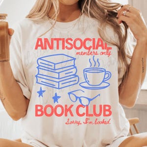 May include: Cream-colored t-shirt with a graphic design. The design features the words "ANTISOCIAL members only BOOK CLUB" in red and blue, along with illustrations of books, a coffee cup, and glasses. The phrase "Sorry, I'm booked" is also included.