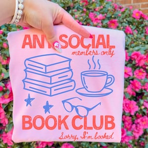 May include: A pale pink sweatshirt featuring the text "ANTI SOCIAL members only BOOK CLUB" in coral. The design includes blue illustrations of books, a coffee cup, stars, and glasses. The phrase "Sorry, I'm booked" is also included.