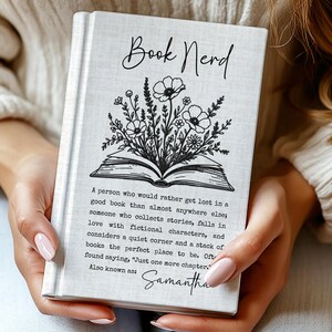 May include: A white hardcover book featuring a black line art illustration of an open book with flowers growing from the pages. The text reads Book Nerd, a definition of a book nerd, and the name Samantha. The cover has a subtle fabric texture.
