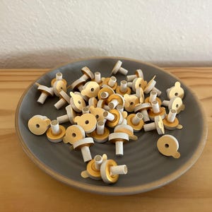May include: A collection of small, mushroom-shaped objects with a white stem, a yellow-gold cap, and a circular base. The objects are scattered on a gray ceramic plate, set against a light-colored wooden surface.