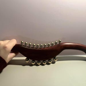 May include: A dark wood massage tool with a curved handle and a series of gold-colored spheres. The tool has a smooth, polished finish and is designed for therapeutic use. The spheres are evenly spaced.