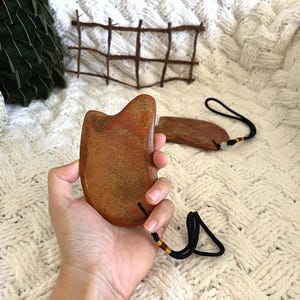 May include: A brown Gua Sha massage tool with a black cord and a small wooden piece. The tool is held in a hand against a white, textured background. The tool is made of stone and has a smooth, curved shape.